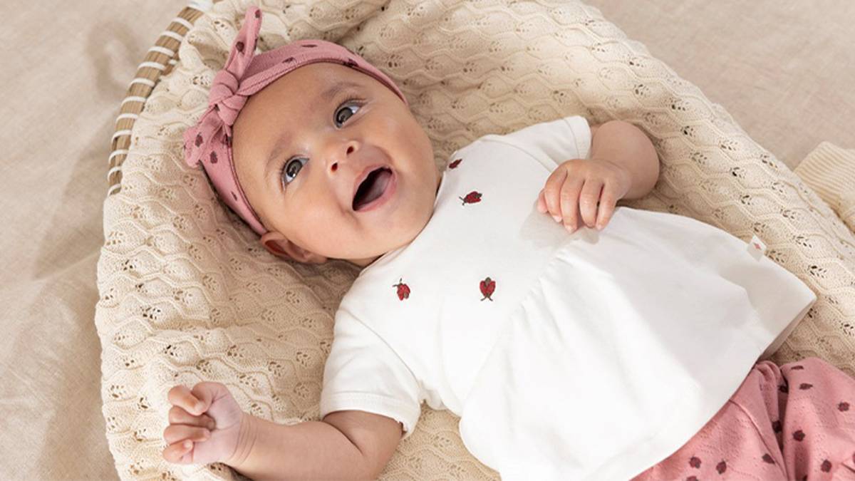 Ein lächelndes Baby mit rosa Stirnband und gemusterter Kleidung liegt in einer gemütlichen Umgebung auf einer weichen Strickdecke.