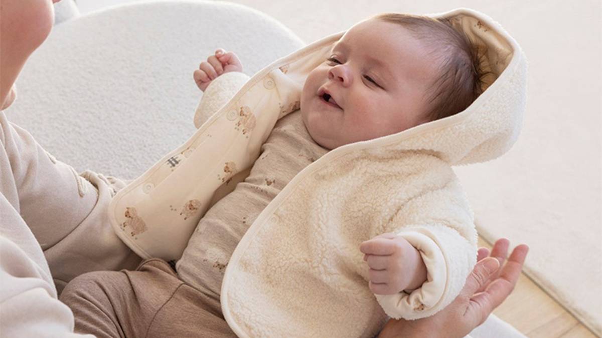 Ein Baby in einer kuscheligen, cremefarbenen Jacke mit Kapuze wird von einem Erwachsenen gehalten, der sanft lächelt. Das Baby trägt helle Kleidung mit einem dezenten Muster.