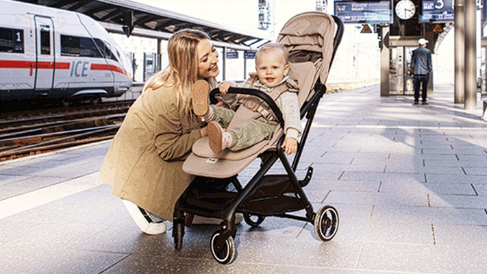 Eine Frau kniet neben einem Baby im Kinderwagen auf einem Bahnsteig; im Hintergrund ist ein ICE-Zug zu sehen.
