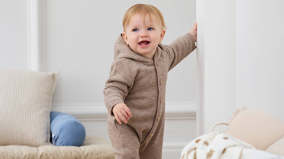 Ein lächelndes Baby in einem braunen Strampler hält sich in einem gemütlichen, sanft beleuchteten Zimmer an einem Türrahmen fest. Kissen und eine Decke liegen in der Nähe.