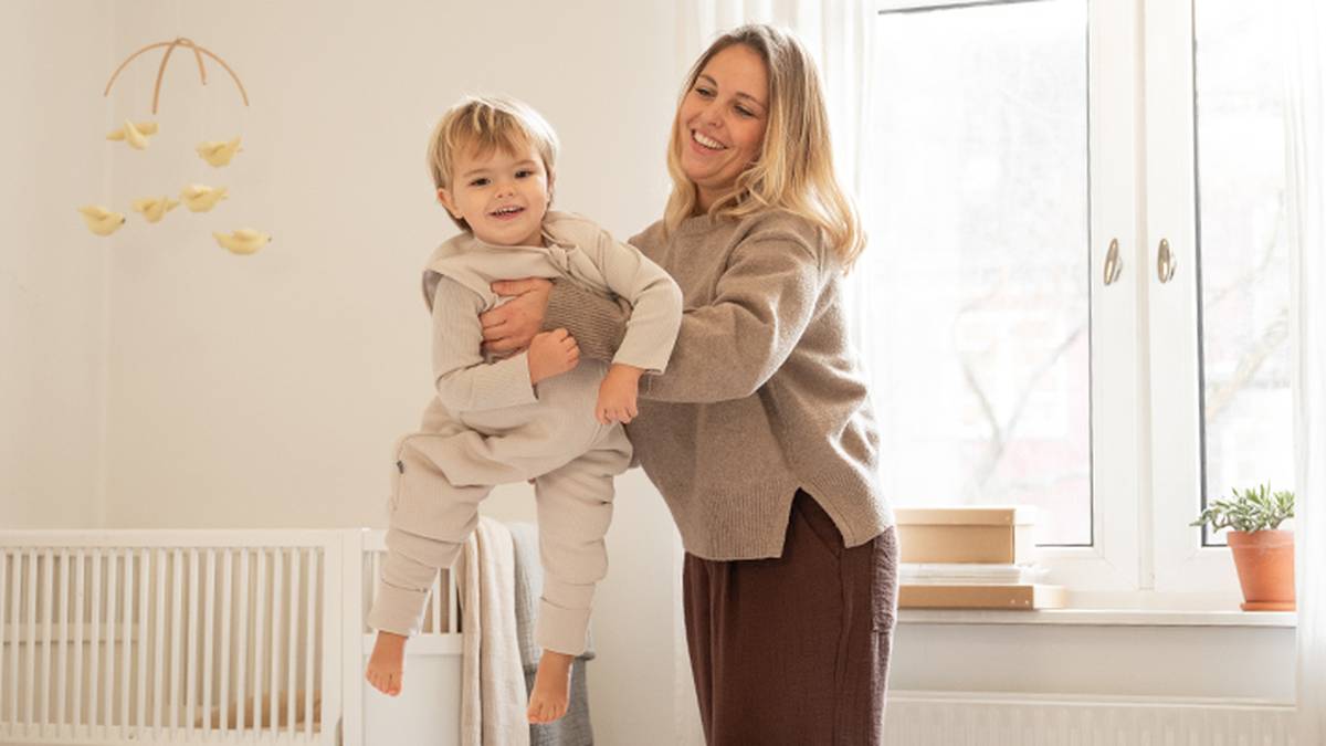 Eine Frau hebt freudig ein lächelndes Kleinkind in einem gemütlichen, hellen Zimmer mit Kinderbett, Mobile und Fenster hoch.