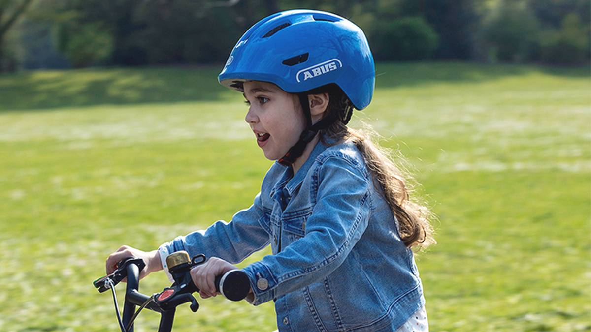Ein Kind mit blauem Helm und Jeansjacke fährt lächelnd und nach vorn blickend Fahrrad in einem grasbewachsenen Park.