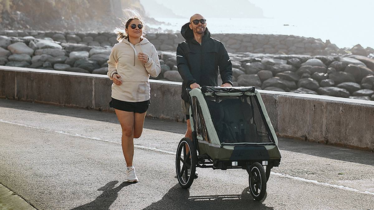 Ein Mann und eine Frau joggen an einem sonnigen Tag mit einem Kinderwagen am Meer entlang, im Hintergrund ist eine felsige Küste zu sehen.
