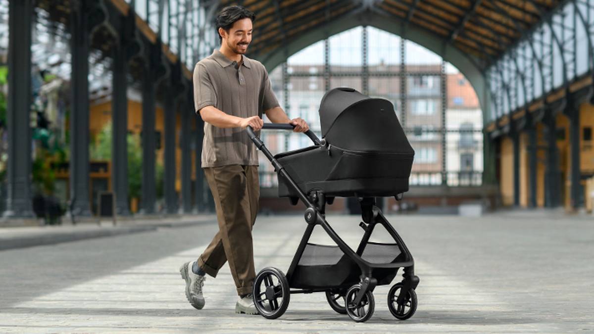 An einem sonnigen Tag geht ein Mann unter einem überdachten Bauwerk hindurch und schiebt lächelnd einen modernen schwarzen Kinderwagen.