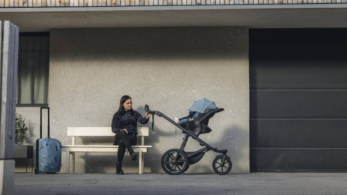 Frau sitzt neben einem Kinderwagen und einem Koffer auf einer Bank vor einem modernen Gebäude mit einem großen Garagentor.