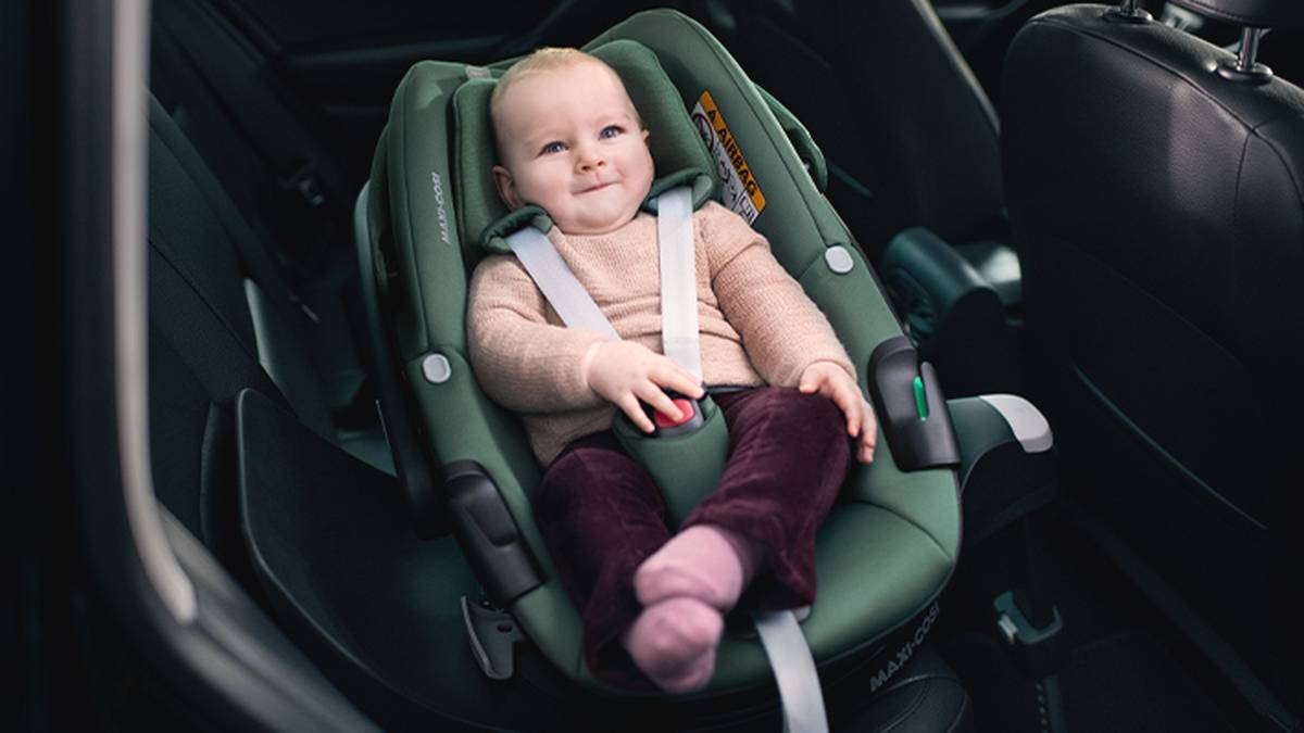 Ein Baby sitzt in einem grünen Kindersitz, trägt einen beigen Pullover und rosa Socken und lächelt im Auto.