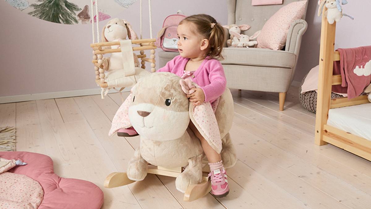 Kleinkindmädchen in rosa Pullover reitet auf einem großen Plüsch-Schaukelhasen in einem pastellfarbenen Kinderzimmer.