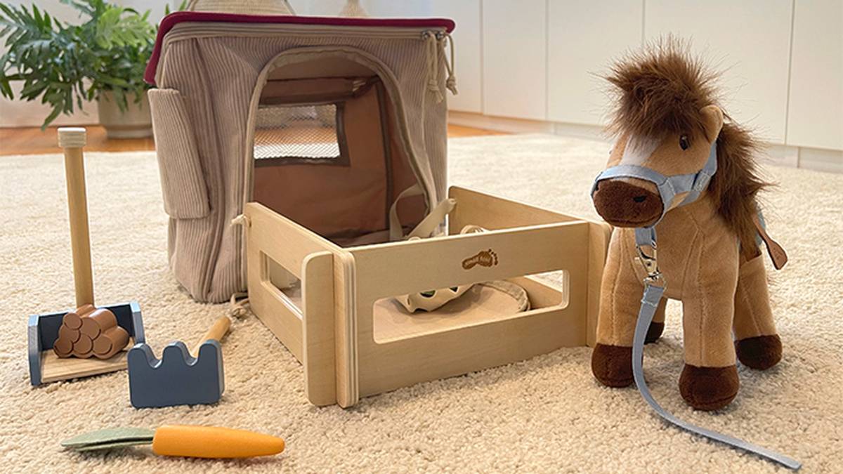 Spielzeugpferd mit Zaumzeug neben einem kleinen Holzstall, einer Plüschtragetasche und landwirtschaftlichen Geräten wie Rechen, Schaufel und Eiern auf einem beigen Teppich.