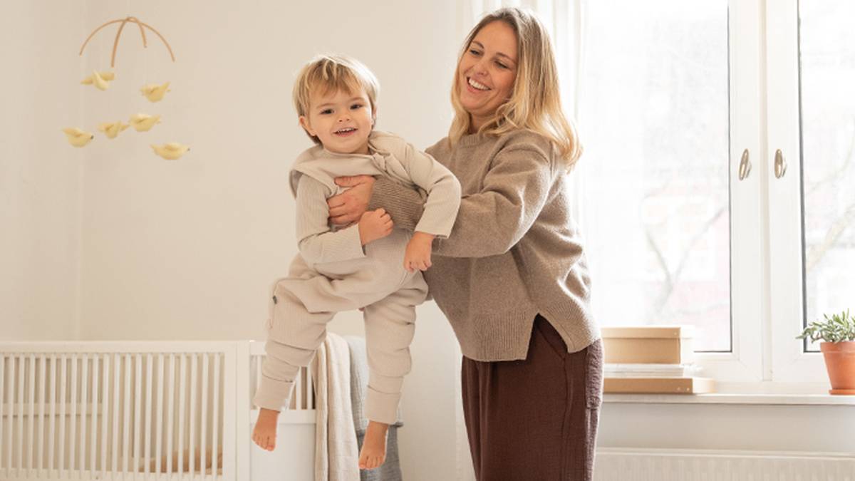 Eine lächelnde Frau hebt ein glückliches Kind in einem gemütlichen, lichtdurchfluteten Zimmer mit Kinderbett, Fenster und Pflanze hoch.