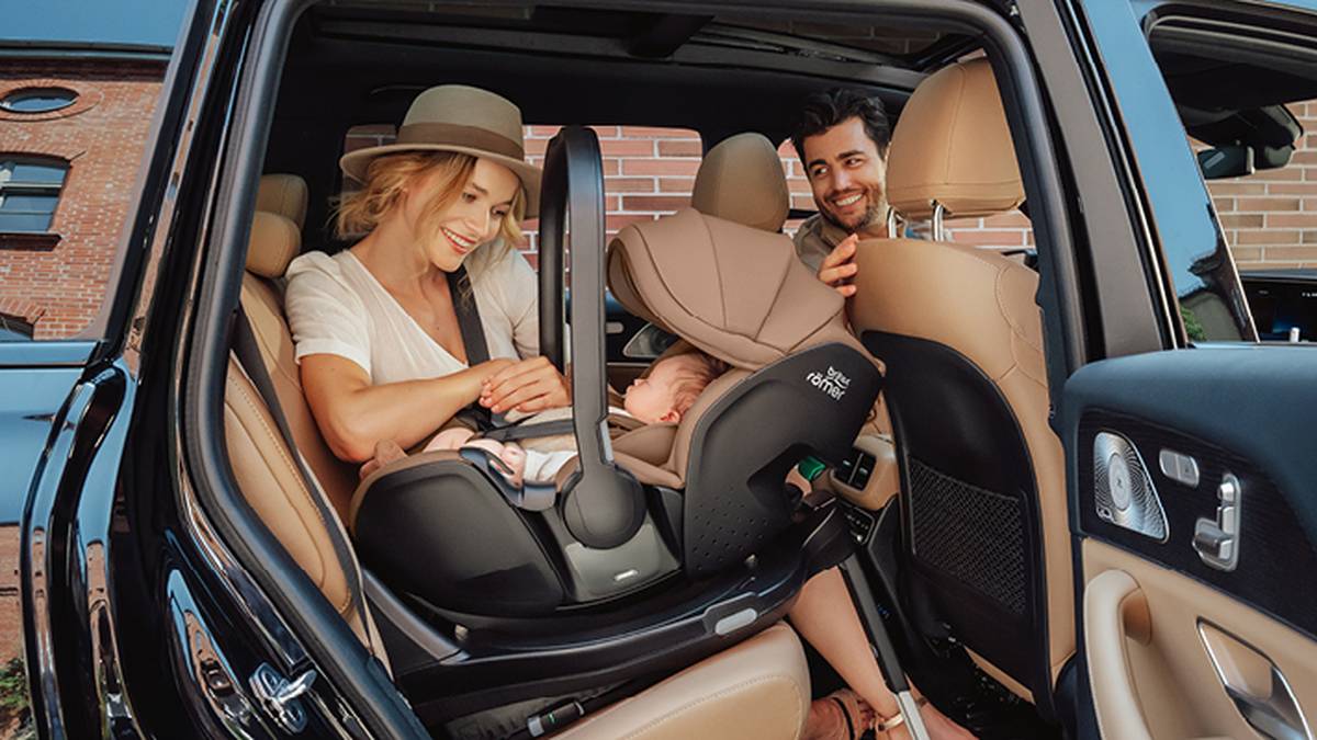 Ein lächelndes Paar sichert ein Baby in einem Autositz in einem geparkten Auto. Die Frau trägt einen Hut und der Mann beugt sich von der Fahrerseite her vor.