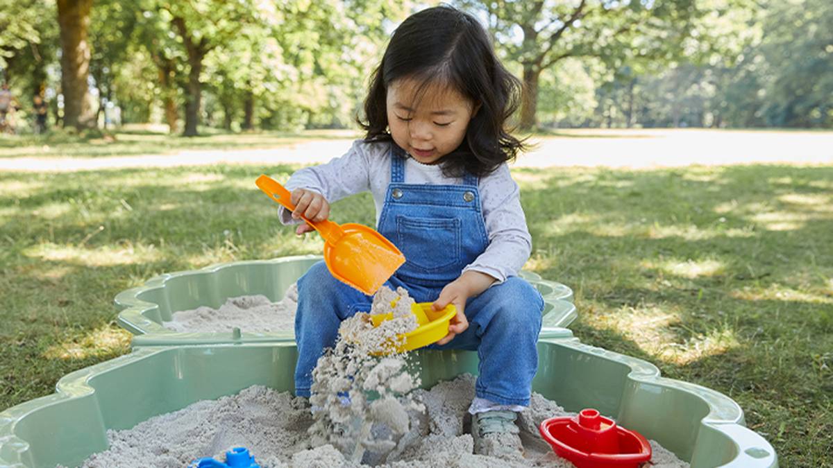 Ein Kind in Jeanslatzhose spielt in einem Sandkasten und schaufelt mit einer gelben Schaufel Sand in einen Eimer. Im Hintergrund sind Bäume und Gras zu sehen.