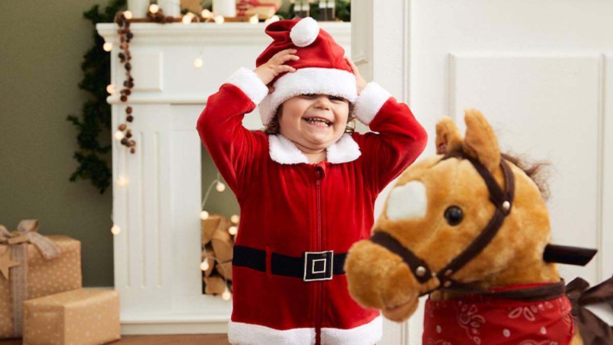 Ein Kind im Weihnachtsmannkostüm lächelt und rückt seine Mütze neben einem Spielzeugpferd zurecht; im Hintergrund sind ein festlich geschmückter Kamin und verpackte Geschenke zu sehen.
