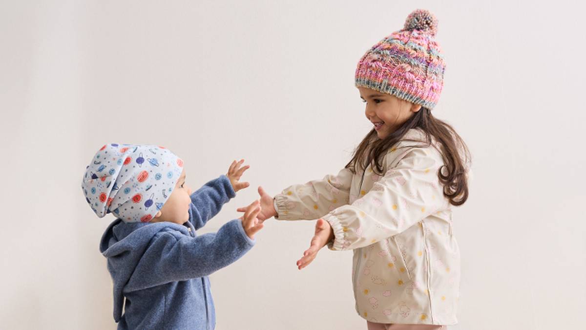 Ein junges Mädchen und ein Kleinkind mit bunten Strickmützen reichen sich vor einem schlichten Hintergrund freudig die Hände.