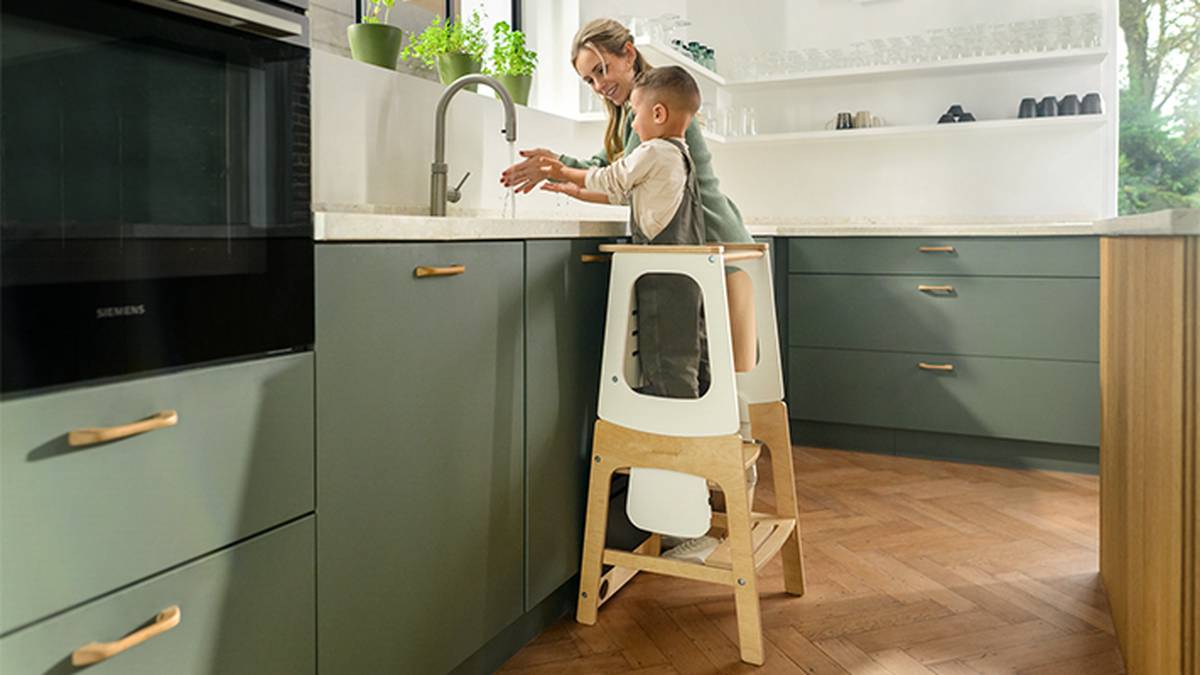 Eine Frau hilft einem Kind beim Händewaschen an der Küchenspüle. Das Kind steht auf einem hölzernen Trittschemel. Die Küche hat grüne Schränke und einen Holzboden.