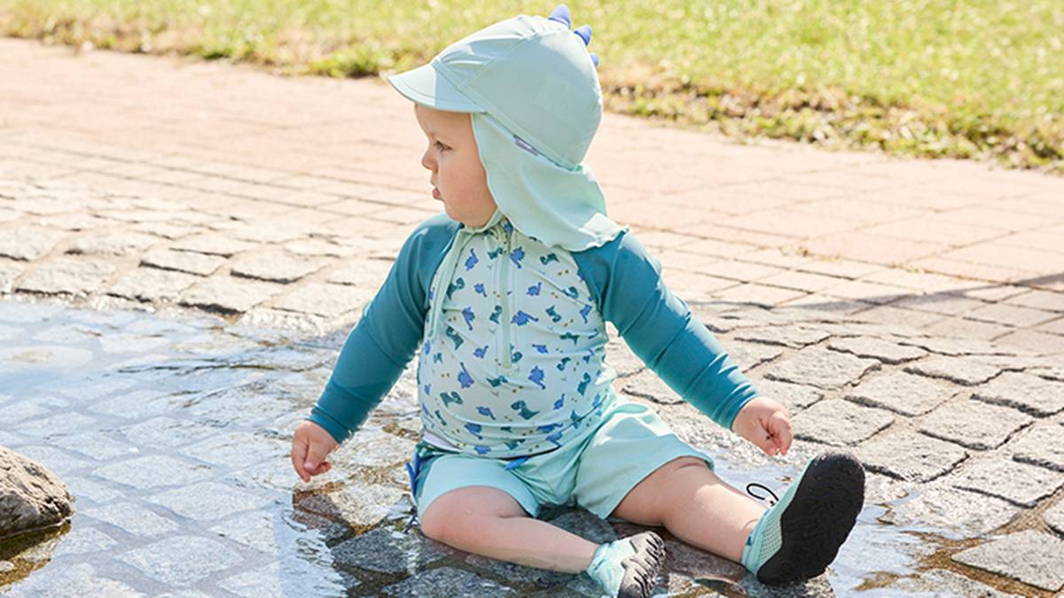 Ein Baby in Badekleidung mit Dinosaurier-Motiv sitzt mit einem Sonnenhut auf dem Kopf im seichten Wasser auf Kopfsteinpflaster, im Hintergrund sind Gras und ein Gehweg zu sehen.