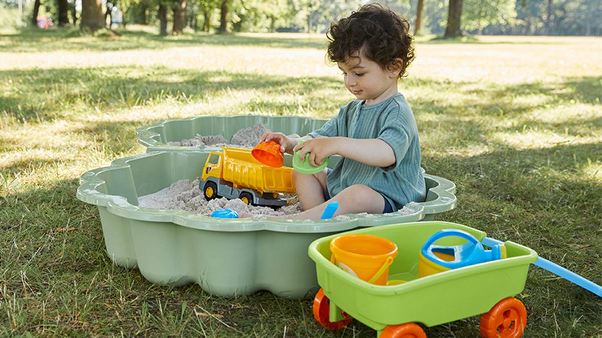 Ein Kind spielt mit einem Spielzeuglaster in einem Sandkasten, umgeben von Bäumen. Neben dem Sandkasten steht ein grüner Bollerwagen mit Sandspielzeug.
