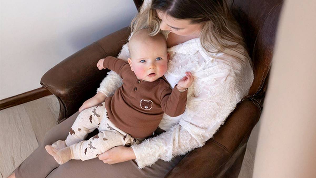 Eine Frau sitzt auf einem braunen Stuhl und wiegt ein Baby in einem braunen Outfit mit Bärenmotiv. Das Baby ruht auf ihrem Schoß und schaut nach oben.