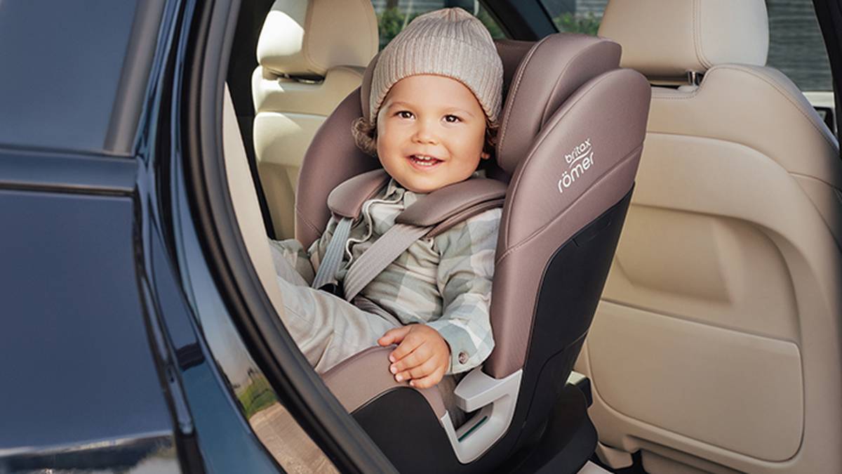 Ein lächelndes Kleinkind sitzt in einem grauen Autositz, trägt eine beige Mütze und ein gestreiftes Outfit und befindet sich in einem Auto mit beiger Innenausstattung.
