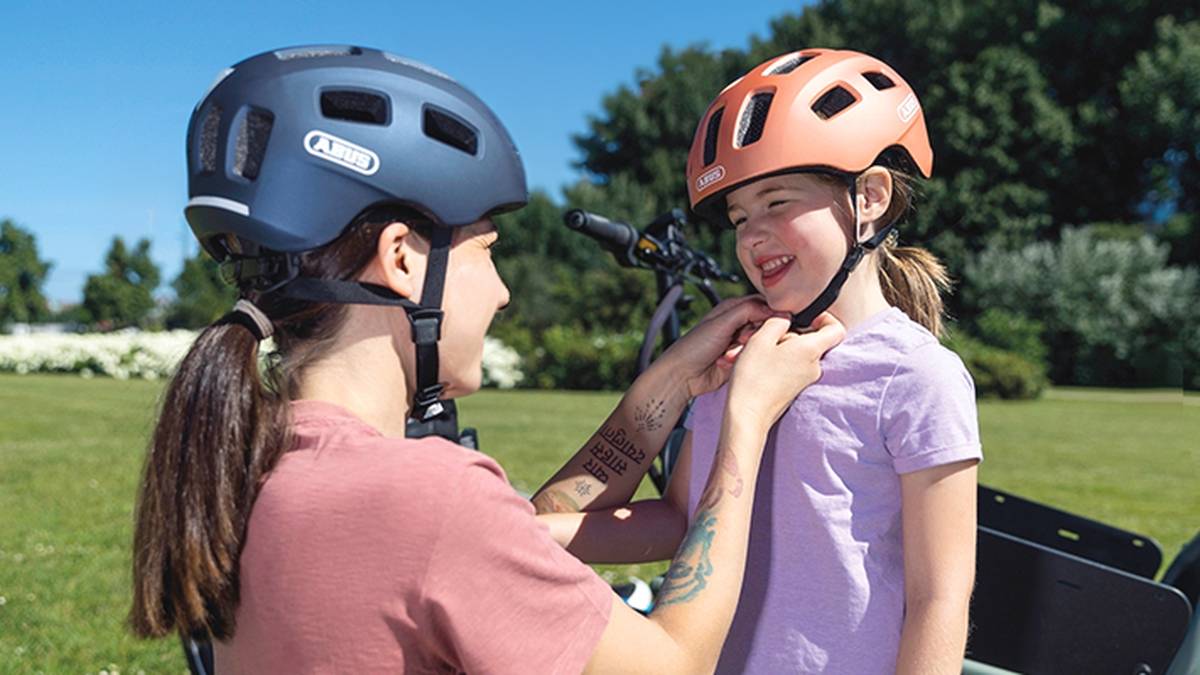 In einem sonnigen Park hilft eine Erwachsene einem Kind, ihren Helm zurechtzurücken. Beide lächeln, und im Hintergrund sind Fahrräder zu sehen.