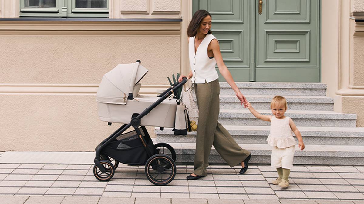 Eine Frau geht mit einem Kleinkind und einem Kinderwagen auf einem gepflasterten Weg vor einem Gebäude mit grünen Türen und Stufen.