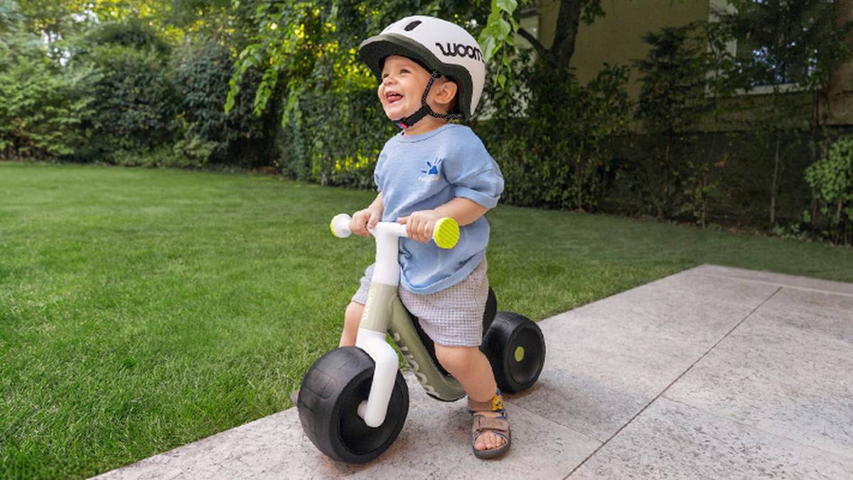 Ein fröhliches Kleinkind mit Helm fährt auf einem Laufrad auf einer Terrasse, im Hintergrund sind grünes Gras und Bäume zu sehen.