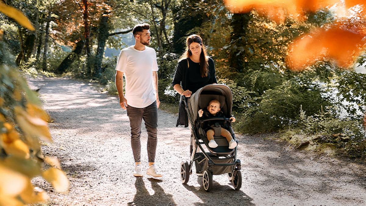 Ein Paar spaziert mit einem Baby im Kinderwagen auf einem sonnenbeschienenen Waldweg, umgeben von üppigem Grün und Herbstlaub.