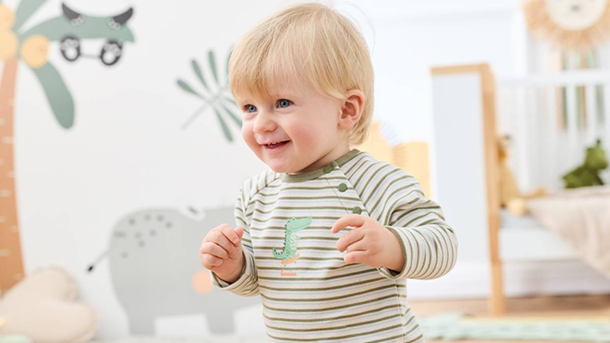 Ein lächelndes Kleinkind in einem gestreiften Strampler mit Dinosaurier-Aufdruck steht in einem Kinderzimmer mit verspielter Wanddekoration und sanfter Beleuchtung.