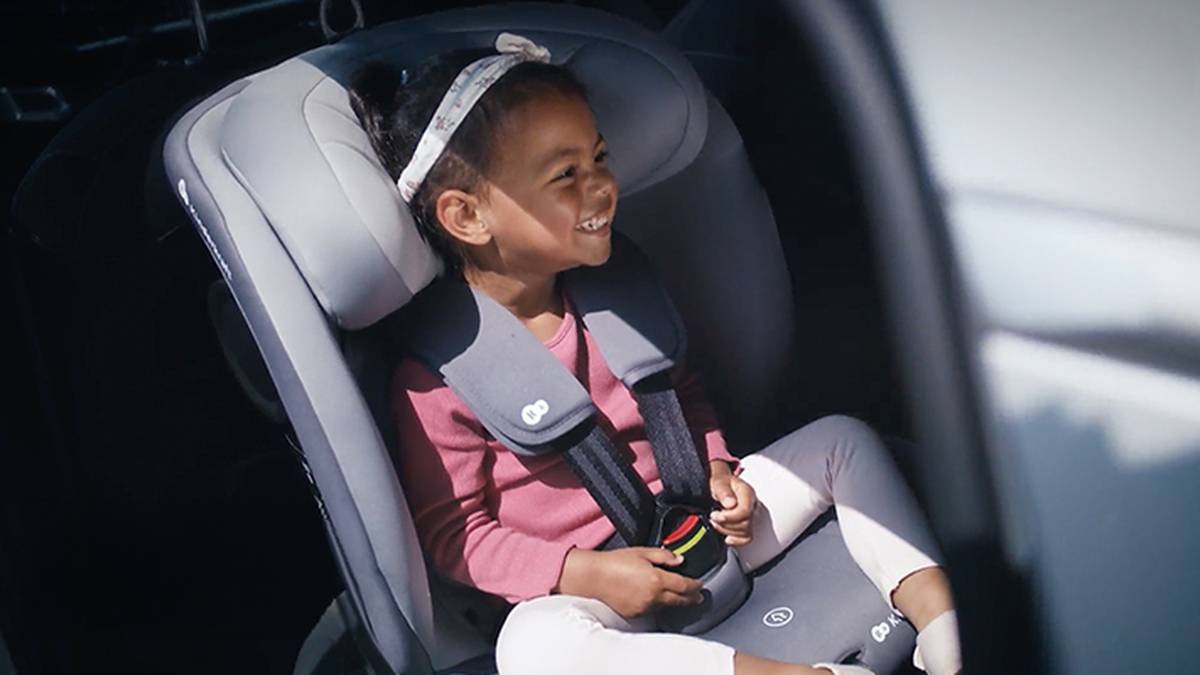 Ein lächelndes Kleinkind in einem rosa Outfit sitzt sicher in einem grauen Kindersitz mit Stirnband im Fahrzeug.