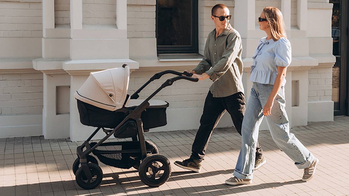 Ein Paar spaziert auf dem Bürgersteig, einer der beiden schiebt einen Kinderwagen. Beide tragen Sonnenbrillen und Freizeitkleidung.