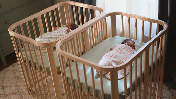 Zwei schlafende Babys, jedes in einem hölzernen Kinderbett, gekleidet in gemusterte Schlafsäcke, durch deren Vorhänge sanftes Tageslicht fällt.