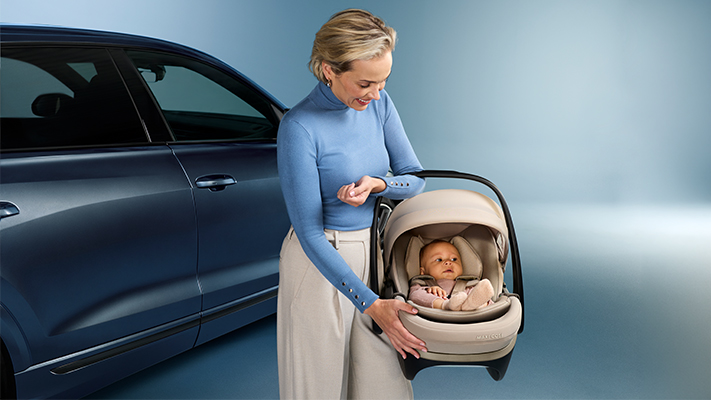 Eine Frau in einem blauen Pullover hält ein Baby in einer Babyschale neben einem dunkelblauen Auto vor einem hellblauen Hintergrund.