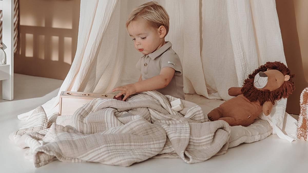 Kleinkind spielt unter einem beigen Baldachin mit Holzspielzeug, sitzt auf einer weichen Decke, neben sich ein Stofflöwe.