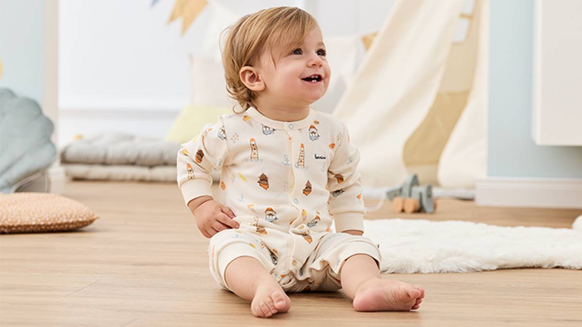 Ein lächelndes Kleinkind in gemustertem Schlafanzug sitzt auf einem Holzboden in einem gemütlichen Zimmer mit Kuscheltieren und heller Einrichtung.