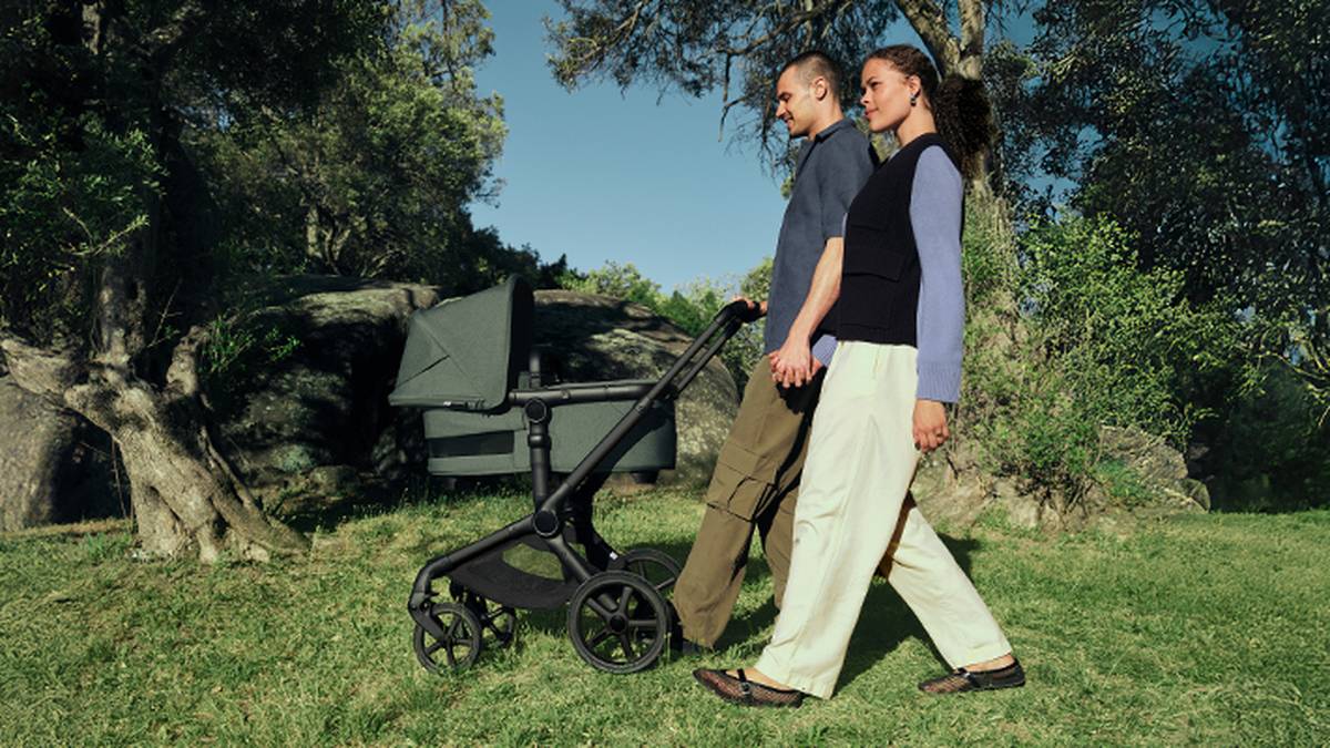 Ein Paar spaziert gemeinsam durch einen Park und schiebt einen grünen Kinderwagen auf einem grasbewachsenen Weg, der von Bäumen und großen Felsen umgeben ist.