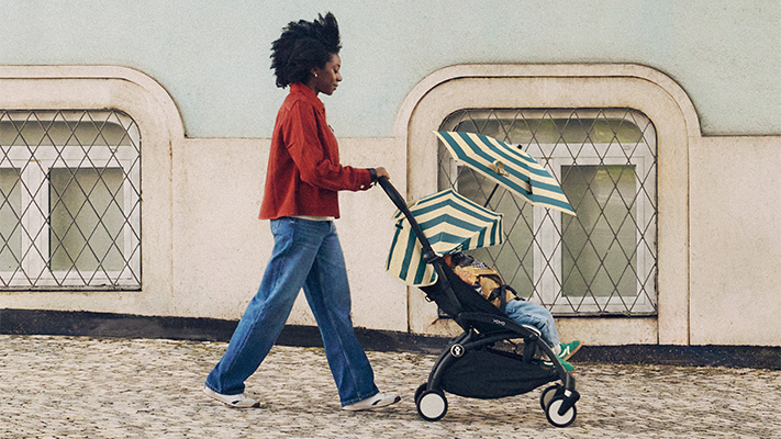 Eine Person in einer roten Jacke schiebt einen Kinderwagen unter einem gestreiften Regenschirm einen Kopfsteinpflasterweg entlang; im Hintergrund sind zwei Bogenfenster zu sehen.