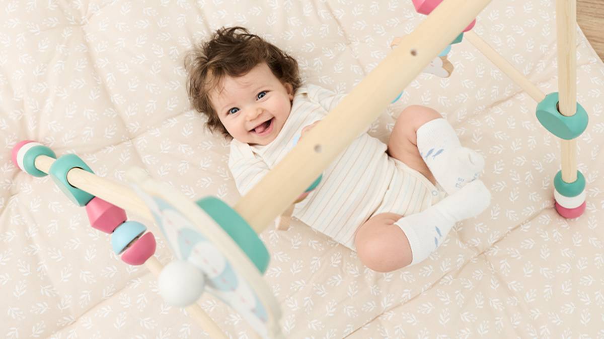 Ein lächelndes Baby in einem gestreiften Strampler liegt auf einer gesteppten Matte und spielt mit einem bunten Babyspielbogen aus Holz.