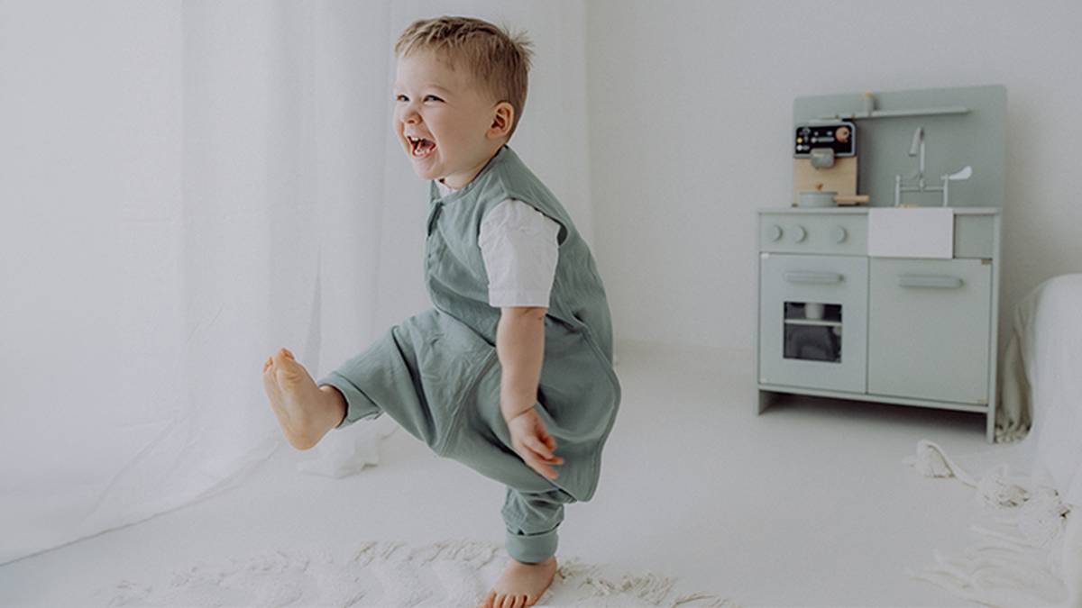 Ein fröhliches Kleinkind in grüner Kleidung lacht und spielt in einem minimalistischen, weißen Zimmer mit einer Spielküche im Hintergrund.