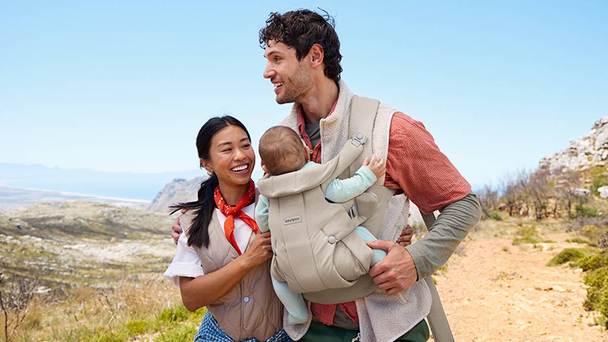 Ein lächelndes Paar spaziert auf einem sonnigen Weg. Der Mann trägt ein Baby in einer hellen Babytrage. Im Hintergrund sind Berge und Himmel zu sehen.