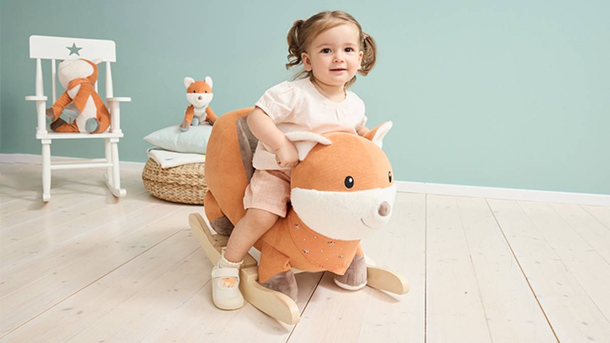 Ein Kleinkind schaukelt in einem hellen Zimmer mit Holzboden auf einem großen, orangefarbenen, fuchsförmigen Schaukeltier. Im Hintergrund sitzt ein passendes Plüschfuchs auf einem Stuhl.