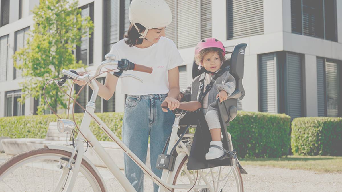 Eine Frau mit Helm schiebt ihr Fahrrad; ein Kind mit rosa Helm sitzt auf einem Fahrradsitz. Im Hintergrund sind moderne Gebäude und Bäume zu sehen.