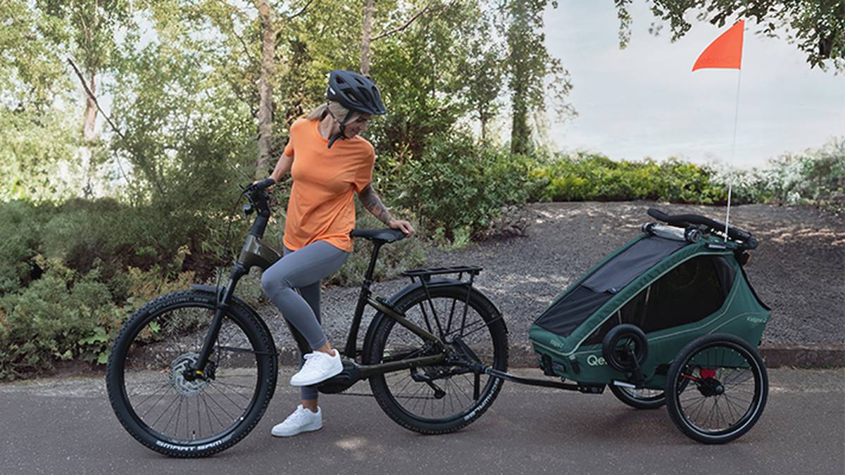Ein Radfahrer in orangefarbenem Hemd und Helm steht mit einem Fahrrad, das an einem grünen Anhänger mit orangefarbener Fahne befestigt ist, auf einem gepflasterten Weg, der von Grün umgeben ist.