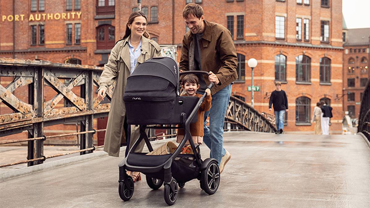 Ein lächelndes Paar schiebt einen Kinderwagen mit einem Kind über eine Brücke, umgeben von Backsteingebäuden und Spaziergängern im Hintergrund.