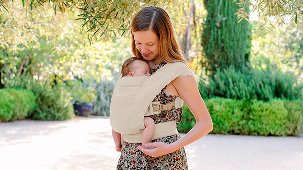 Eine Frau steht im Freien und lächelt auf ein Baby in einer beigen Trage hinab. Üppiges Grün umgibt sie und schafft einen ruhigen Hintergrund.