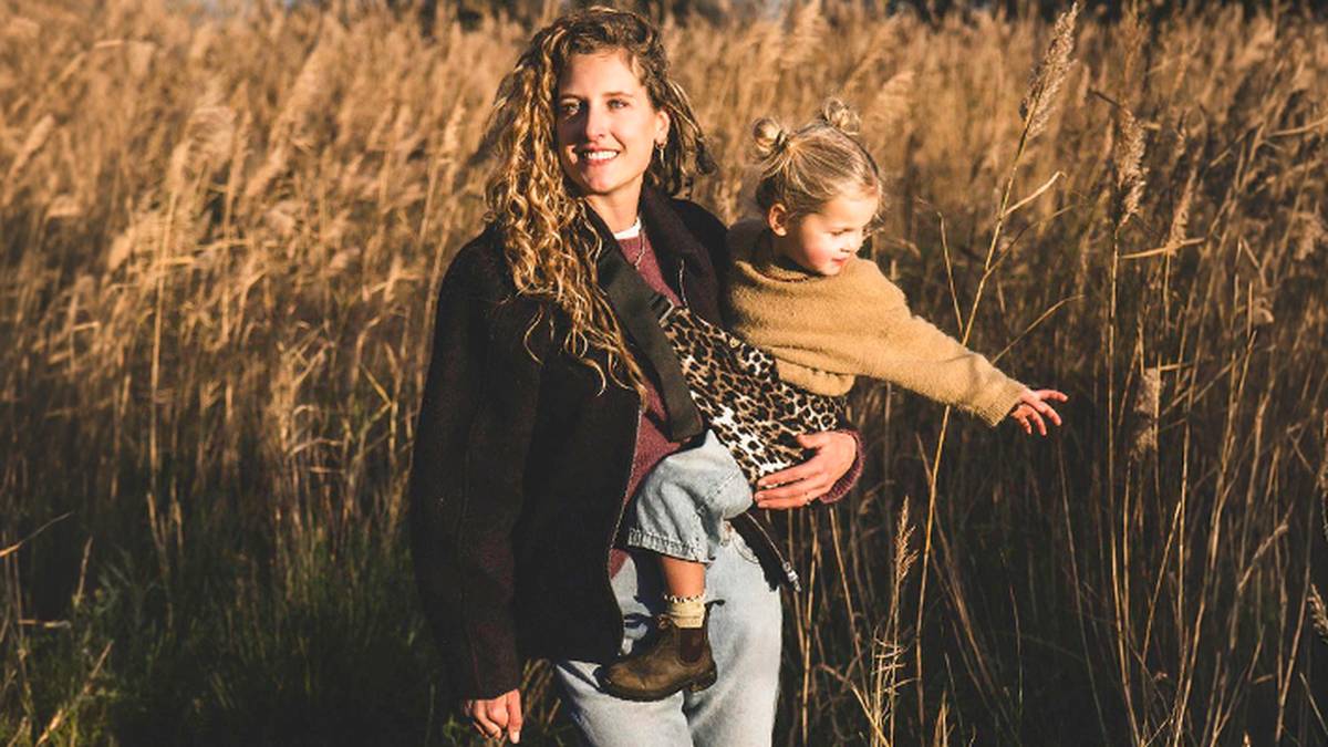 Eine Frau mit langen, lockigen Haaren hält ein kleines Kind, das einen Schal mit Leopardenmuster trägt. Sie stehen in einem Feld mit hohem, goldenem Gras im Sonnenlicht.