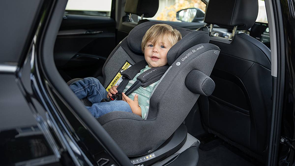 Ein Kleinkind sitzt sicher in einem grauen Autositz auf der Rückbank eines Fahrzeugs, lächelt und trägt ein grünes Hemd und Jeans.