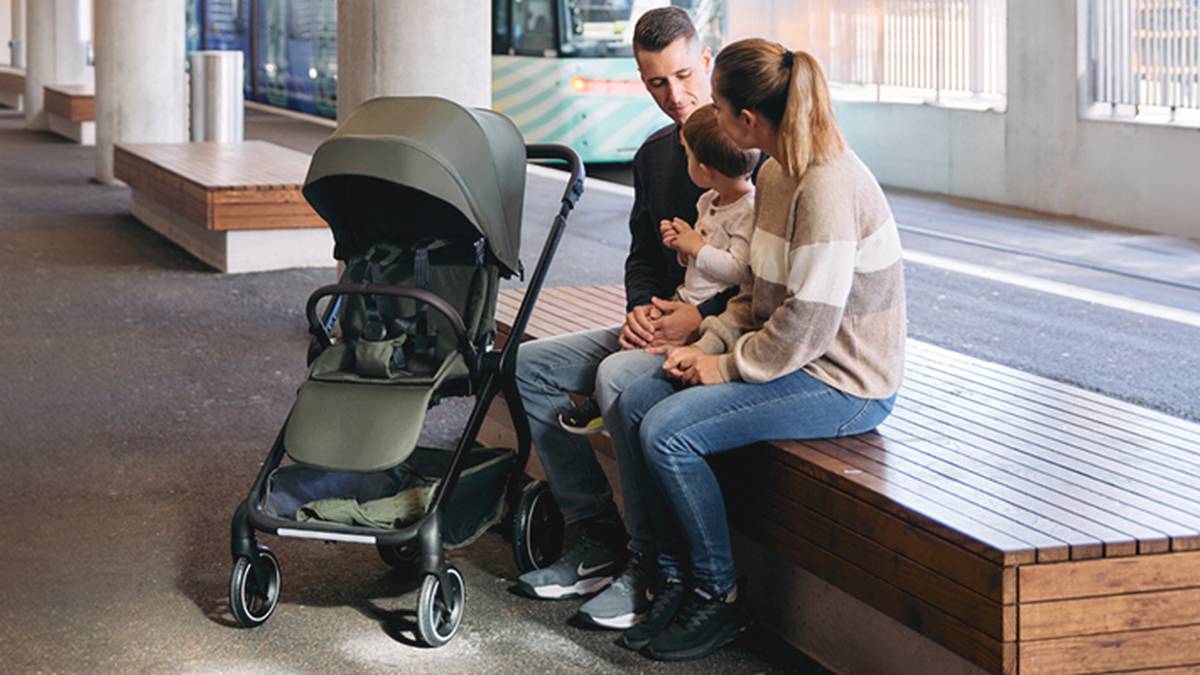 Ein Paar sitzt mit einem Baby auf dem Schoß auf einer Bank neben einem Kinderwagen in einer städtischen Umgebung; im Hintergrund ist eine Straßenbahn zu sehen.