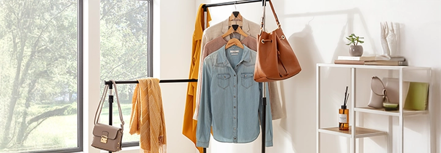 Kleiderständer mit drei verschiedenen Hemden, einem Schal, einem orangefarbenen Mantel und Handtaschen in der Nähe eines Fensters. Regale mit Dekoartikeln und einer Pflanze auf der rechten Seite.