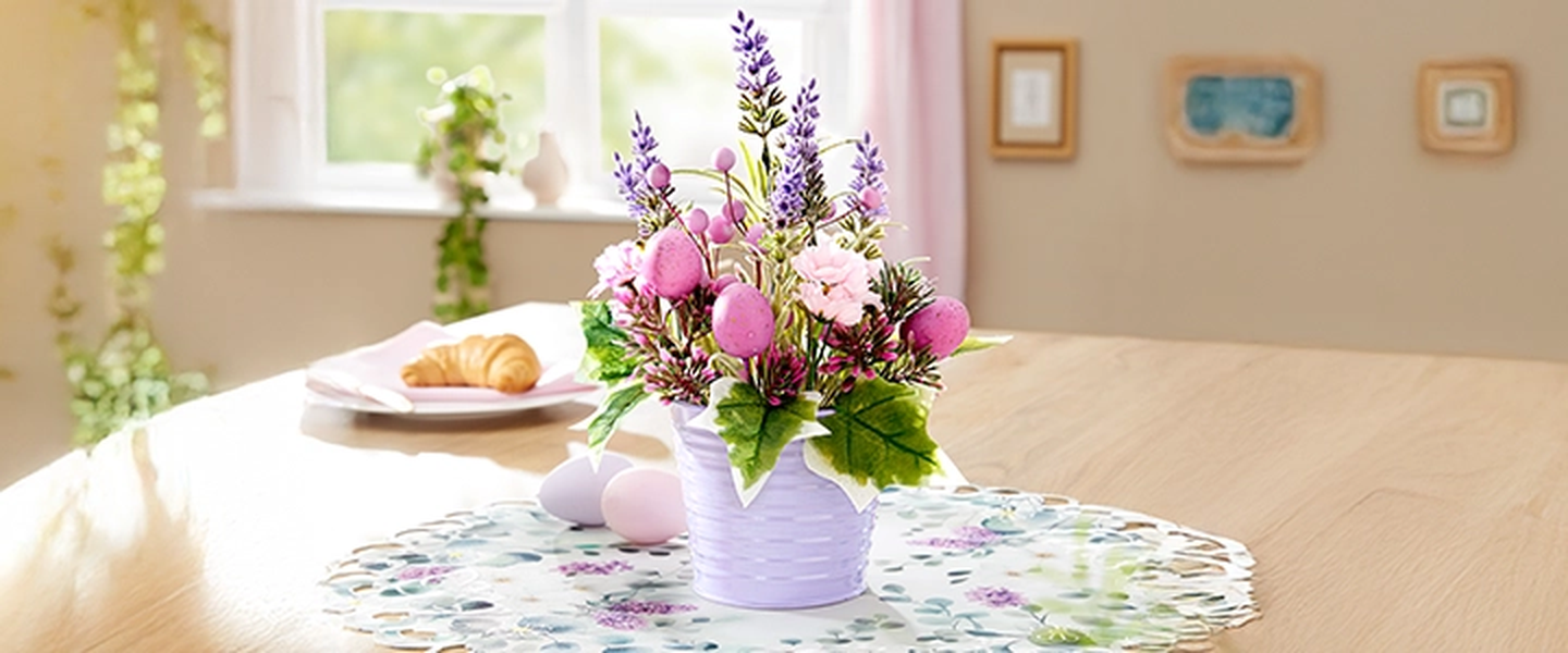 Eine Vase mit rosa und lila Blumen auf einem gemusterten Tischtuch auf einem Holztisch, Sonnenlicht strömt durch ein Fenster und daneben steht ein Teller mit einem Croissant.
