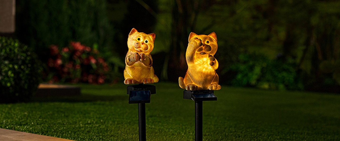 Zwei Solarleuchten in Katzenform auf Erdspießen beleuchten nachts einen Garten, umgeben von Pflanzen und einem dunklen Hintergrund.
