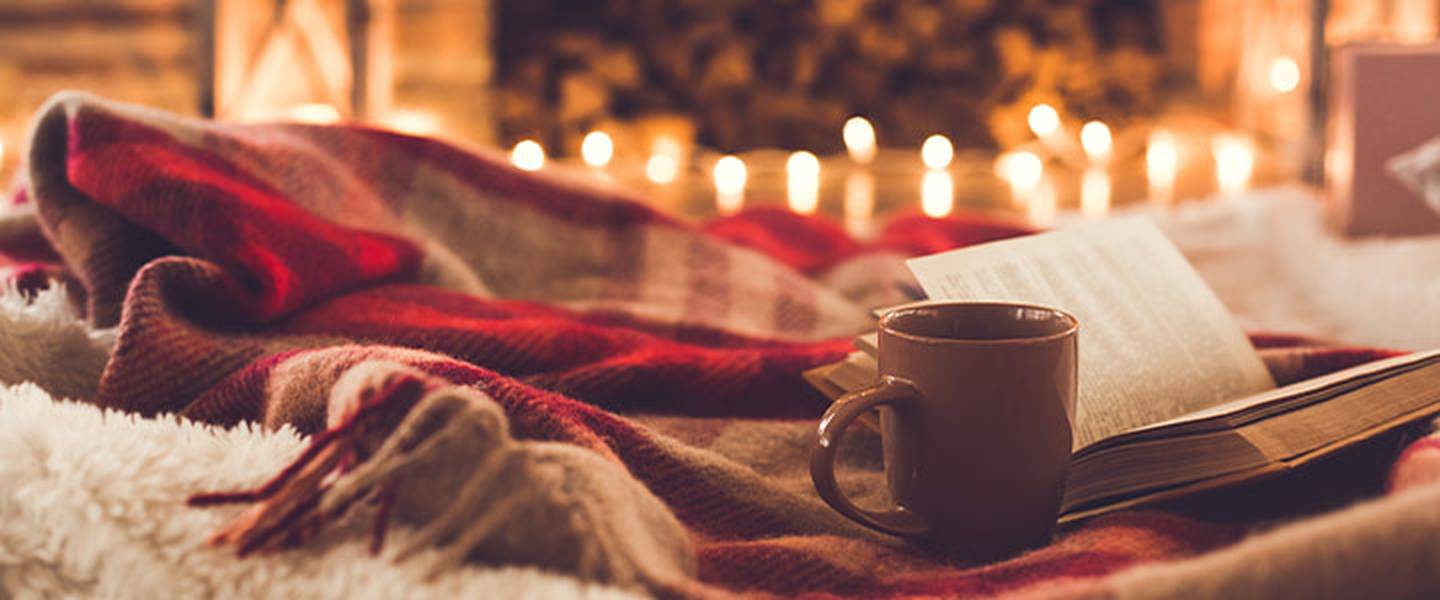 Gemütliche Szene mit einem aufgeschlagenen Buch, einer Tasse und einer rot-karierten Decke. Warme Lichter funkeln im Hintergrund und schaffen eine behagliche Atmosphäre.